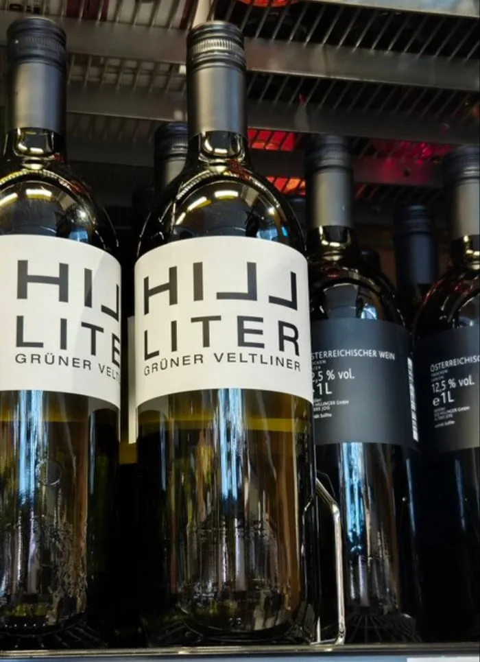 Several wine bottles are lined up on a store shelf, with two prominently featuring a white label that reads 'HIJJ LITER…