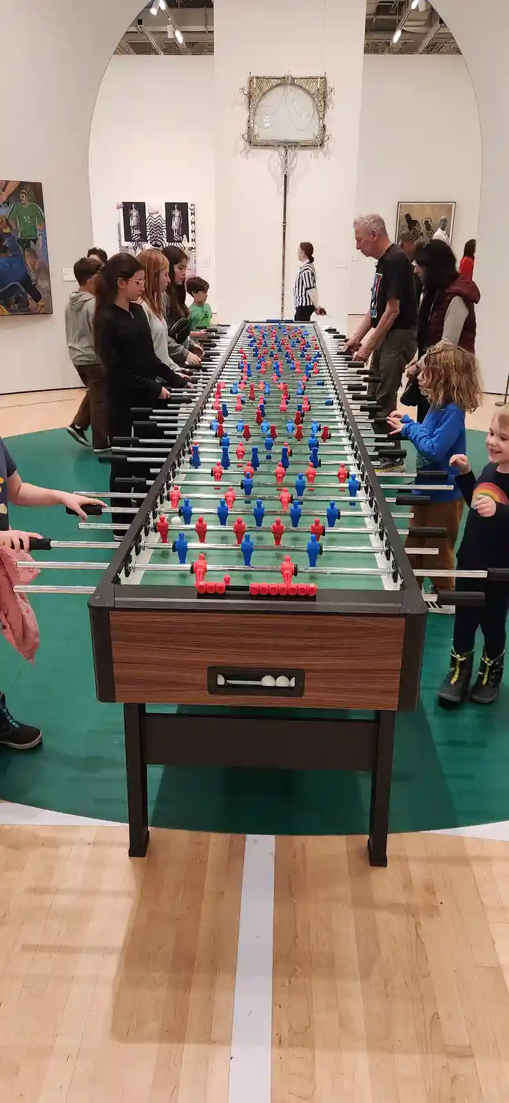A long foosball table is set up in what appears to be a museum gallery, surrounded by people of various ages playing…