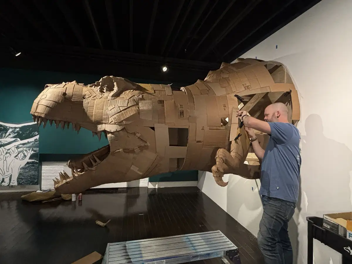 A man in a blue t-shirt is working on a large, intricate cardboard sculpture of a Tyrannosaurus rex in what appears to be a…