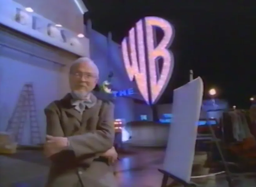 an older man with glasses and a beard stands with arms crossed in front of a large, illuminated 'WB' logo at night, with an…