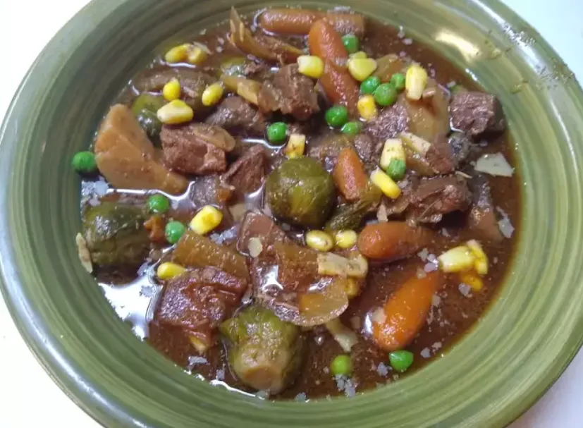 A close-up photo of a green bowl filled with a stew containing chunks of meat, carrots, peas, corn, and brussels sprouts in…