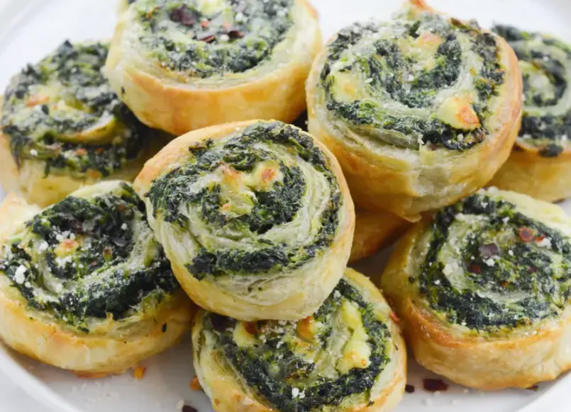 A close-up photo of golden-brown, spiral-shaped pinwheels filled with green spinach and cheese, topped with red pepper…