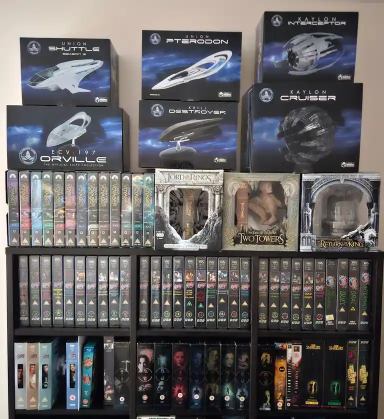 A bookshelf displays a large collection of science fiction and fantasy media, including boxed model kits of fictional…