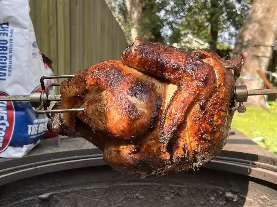 A whole roasted chicken spins on a rotisserie over a grill, its skin golden-brown and charred in places. Behind it, a large…