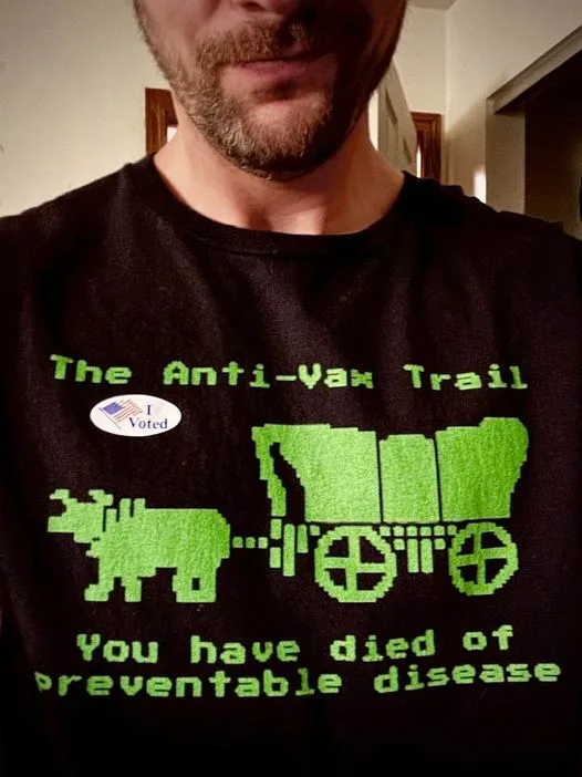 A man wearing a black t-shirt with green pixelated text and graphics. The shirt reads 'The Anti-Vax Trail' above a pixelated…