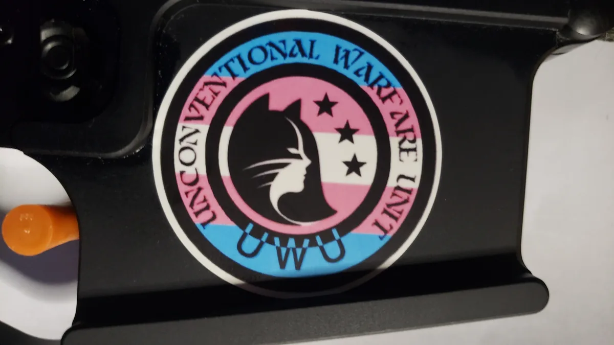 A circular sticker on a black surface features a black cat silhouette with a white outline, set against a pink, white, and…