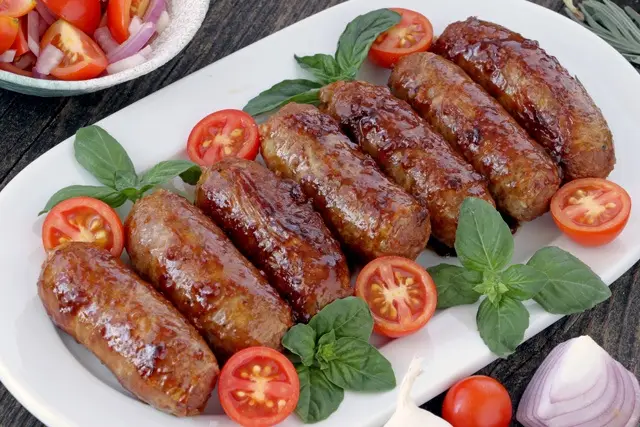 A white platter holds six glossy, grilled sausages arranged in a row, garnished with fresh basil leaves and halved cherry…