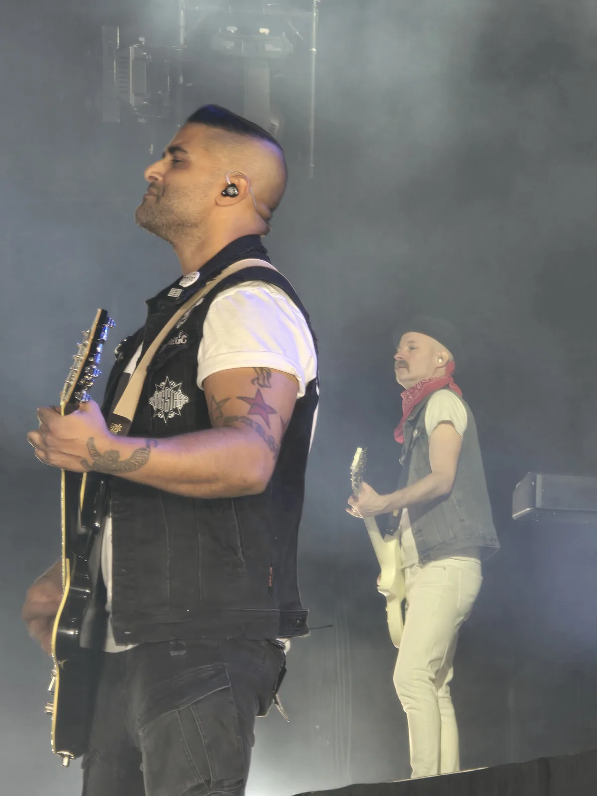 Two male musicians are on stage during a concert, both holding electric guitars and wearing in-ear monitors. The man in the…
