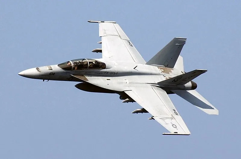 A U.S. Navy F/A-18 Hornet fighter jet is captured in flight against a clear blue sky, banking slightly with its landing gear…