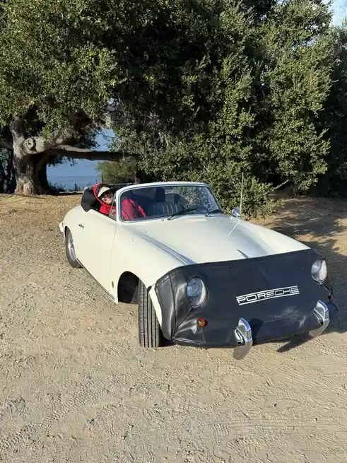 A person in a red jacket and cap sits in the driver's seat of a white vintage Porsche convertible parked on a dirt path…