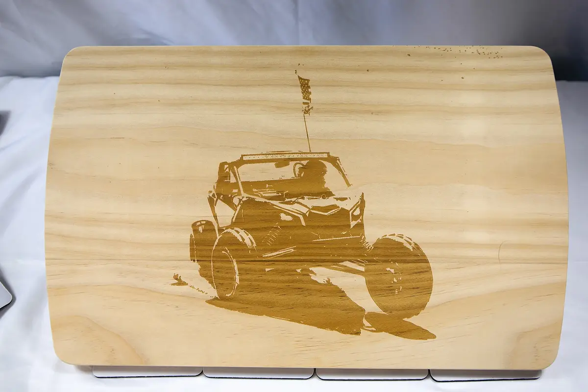 A light-colored wooden cutting board features a laser-etched image of a rugged off-road vehicle, resembling a UTV or dune…