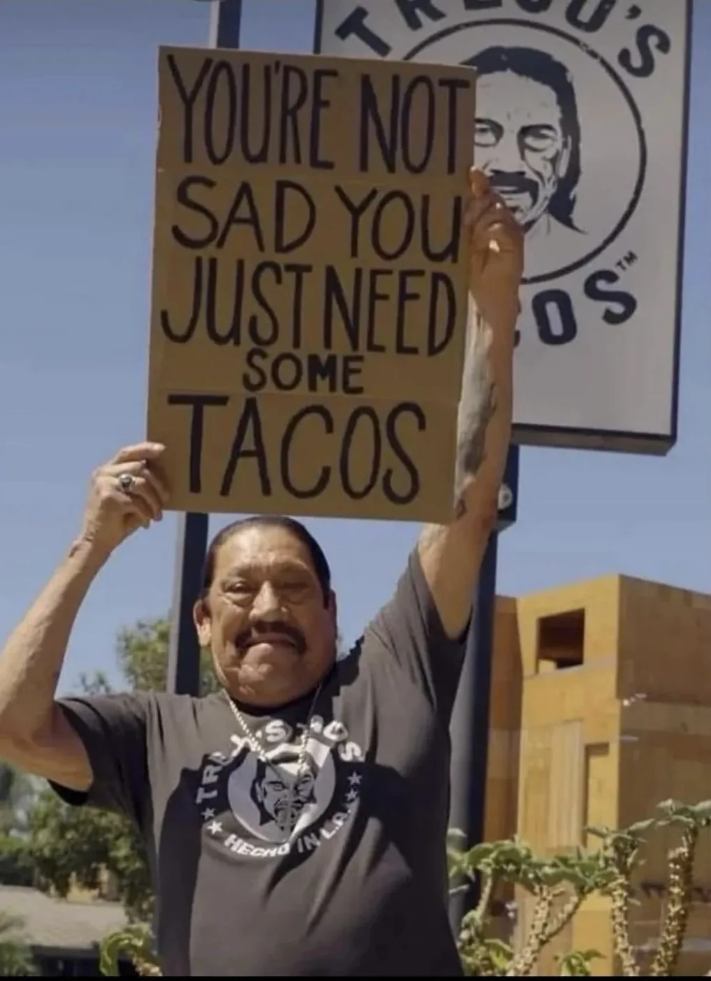 A man in a Tres Tacos branded t-shirt holds up a cardboard sign that reads 'YOU'RE NOT SAD YOU JUST NEED SOME TACOS' in…