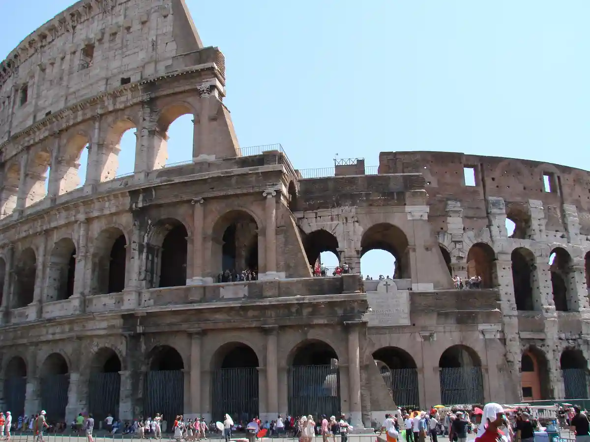 The image shows the ancient Colosseum in Rome, Italy, with its iconic arched structure and weathered stone facade under a…