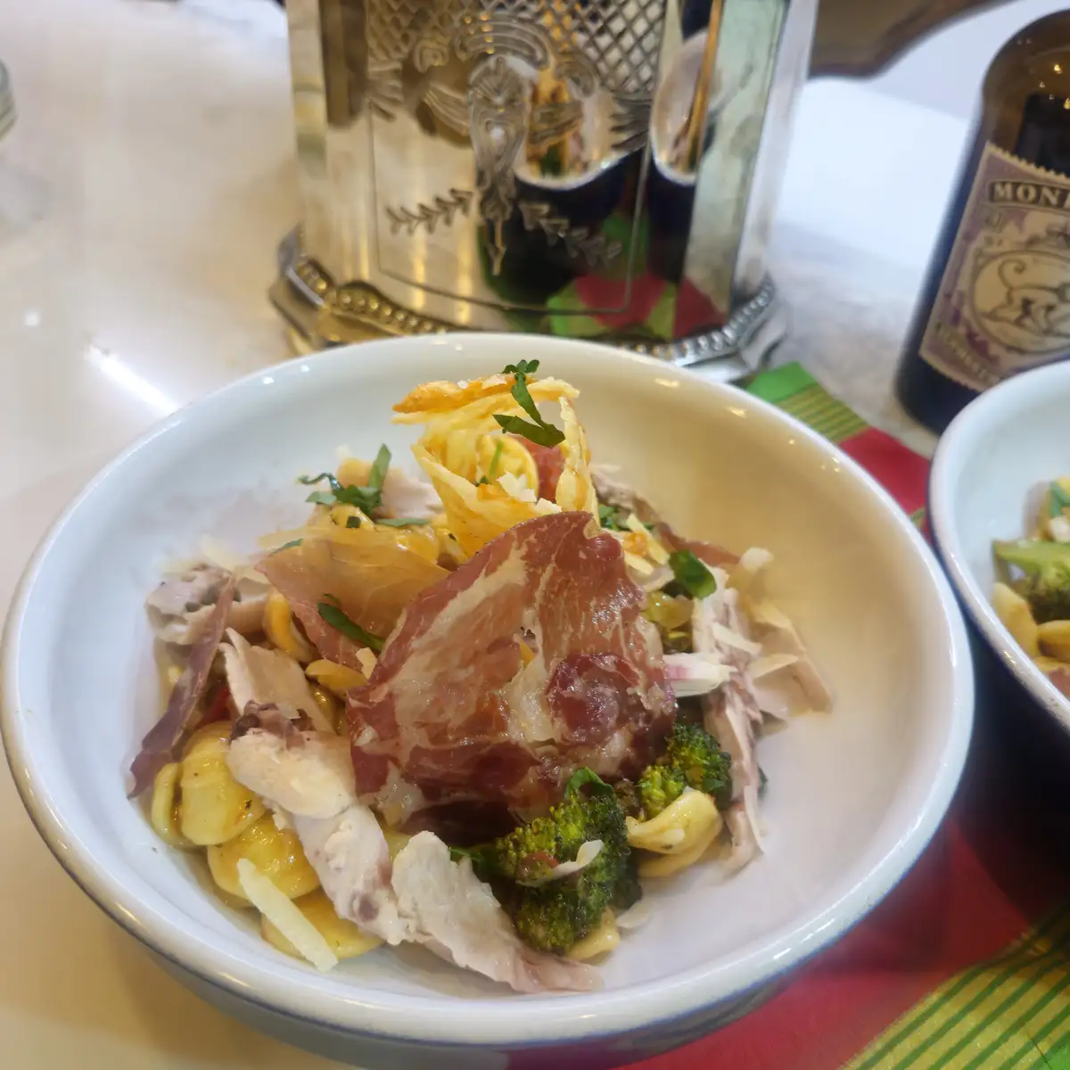 A close-up photo of a white bowl filled with pasta, chicken, broccoli, and a large slice of cured meat, with a decorative…