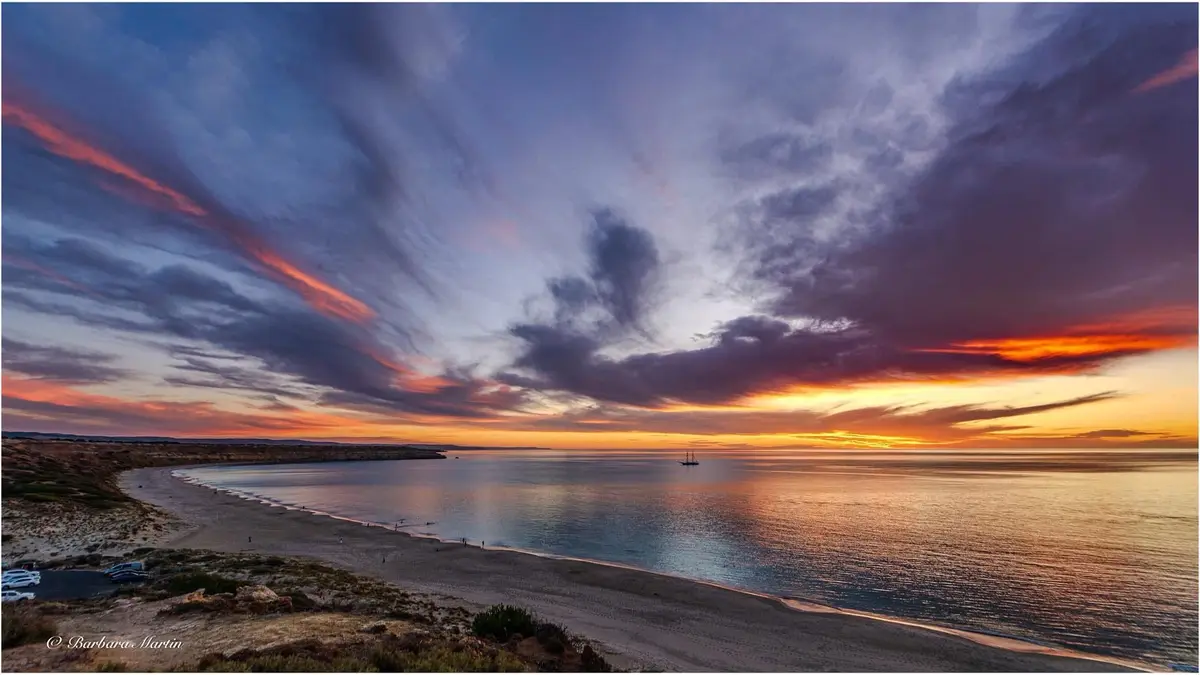 A breathtaking sunset photograph of a calm bay with vibrant orange, purple, and blue clouds reflecting on the water. A lone…