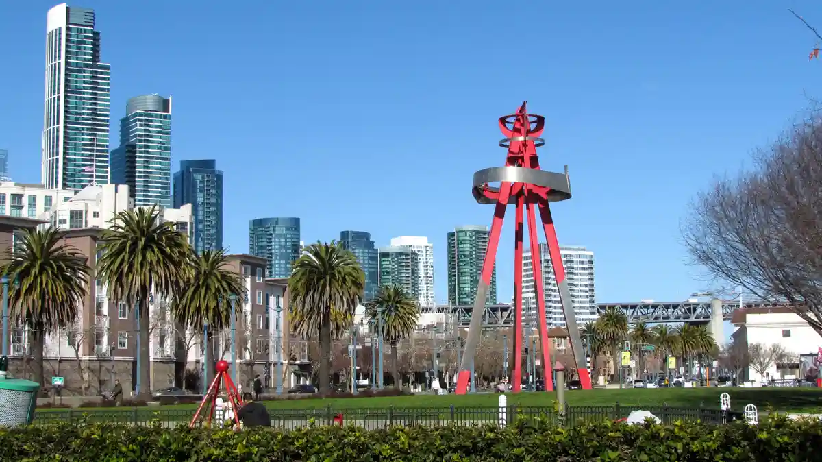 A sunny urban park scene with palm trees, a black fence, and a large red abstract sculpture in the foreground. Behind it, a…