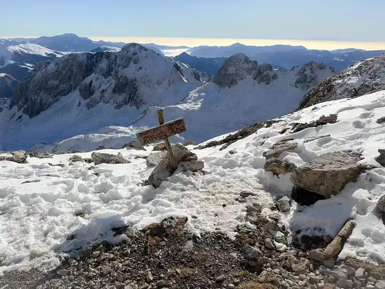 A wooden trail sign in a snowy mountain landscape points right toward 'Rifugio Olmo' at 320°, with a vast panorama of…
