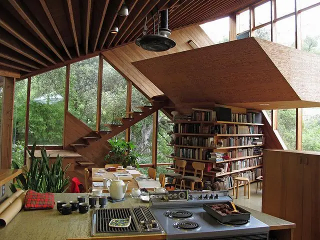 a modern wooden house interior with a kitchen counter, stove, and dining table, but a large bookshelf and staircase are…