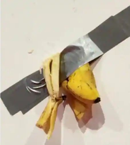 A ripe yellow banana is taped with black duct tape to the blade of a kitchen knife, which is lying on a plain white surface…