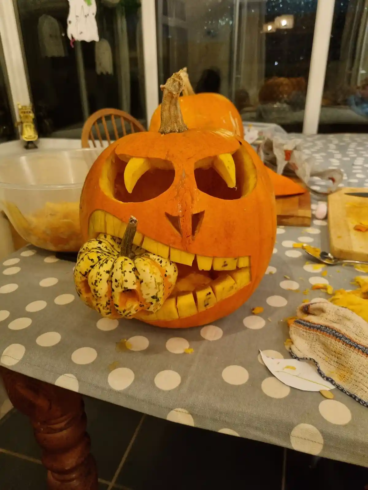 A large orange pumpkin carved with a wide, toothy grin and jagged eyes sits on a polka-dotted tablecloth, holding a smaller…