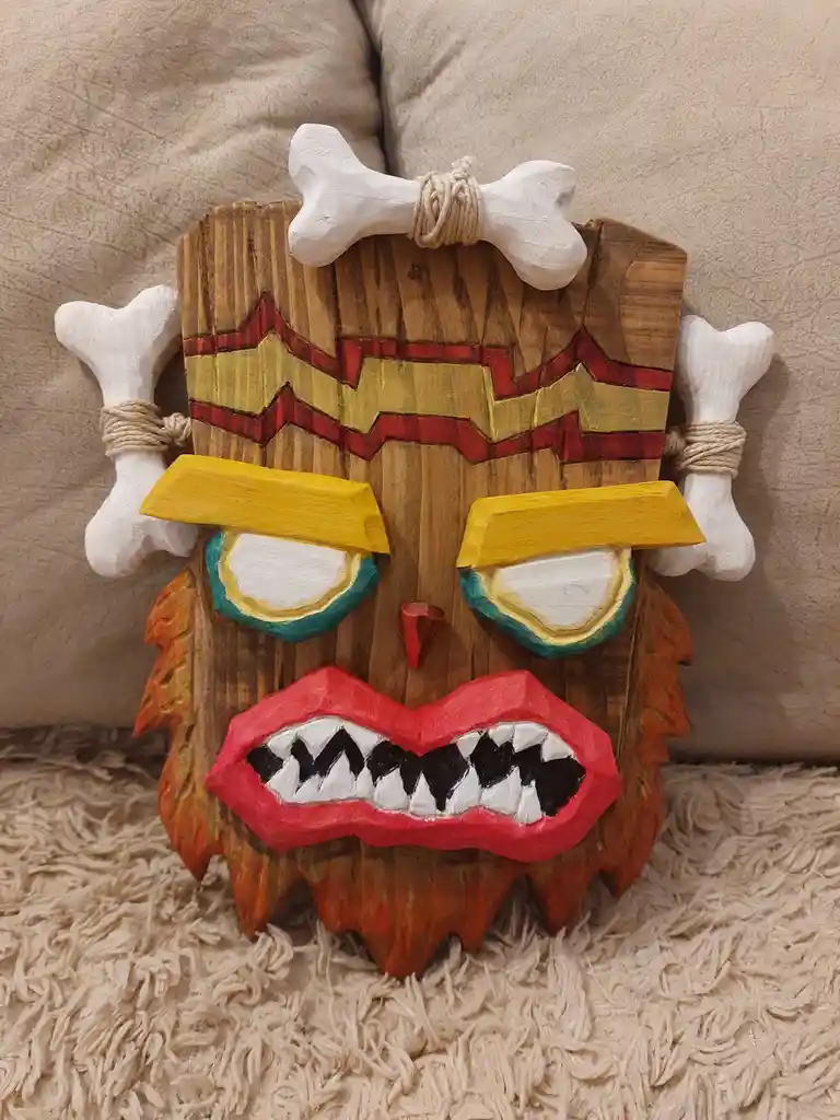 A handcrafted wooden tiki mask with painted red, yellow, and teal features, sharp teeth, and white bone decorations tied…