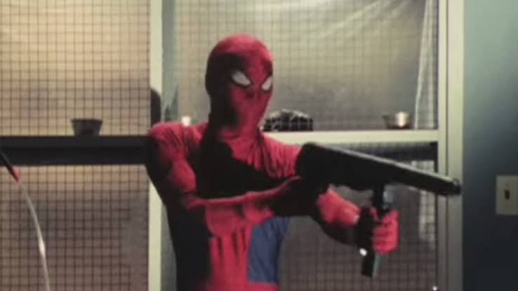 A person in a red and blue Spider-Man costume stands indoors holding a black hairdryer like a weapon, aiming it forward with…