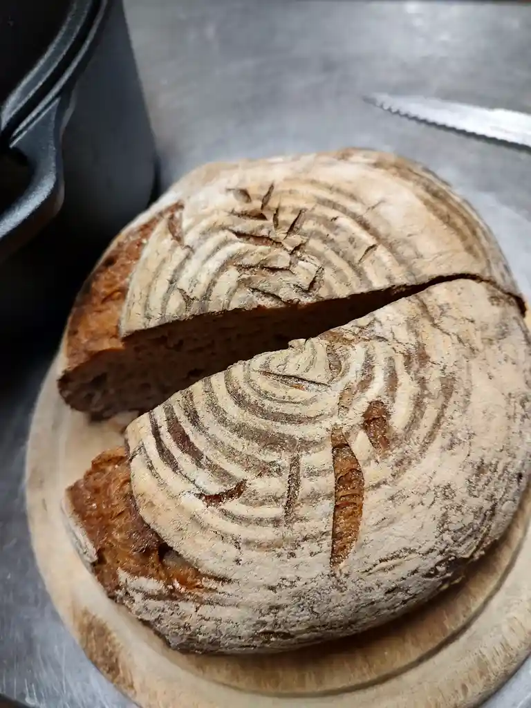 A round loaf of artisan bread with a cracked, textured crust resembling tree bark sits sliced open on a wooden board. The…
