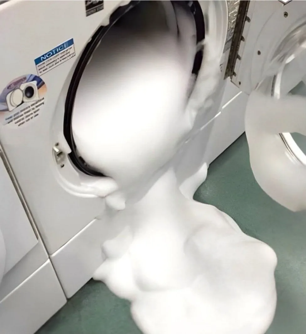 A white front-loading washing machine is open, spilling a large amount of thick, white foam onto the green floor. The foam…