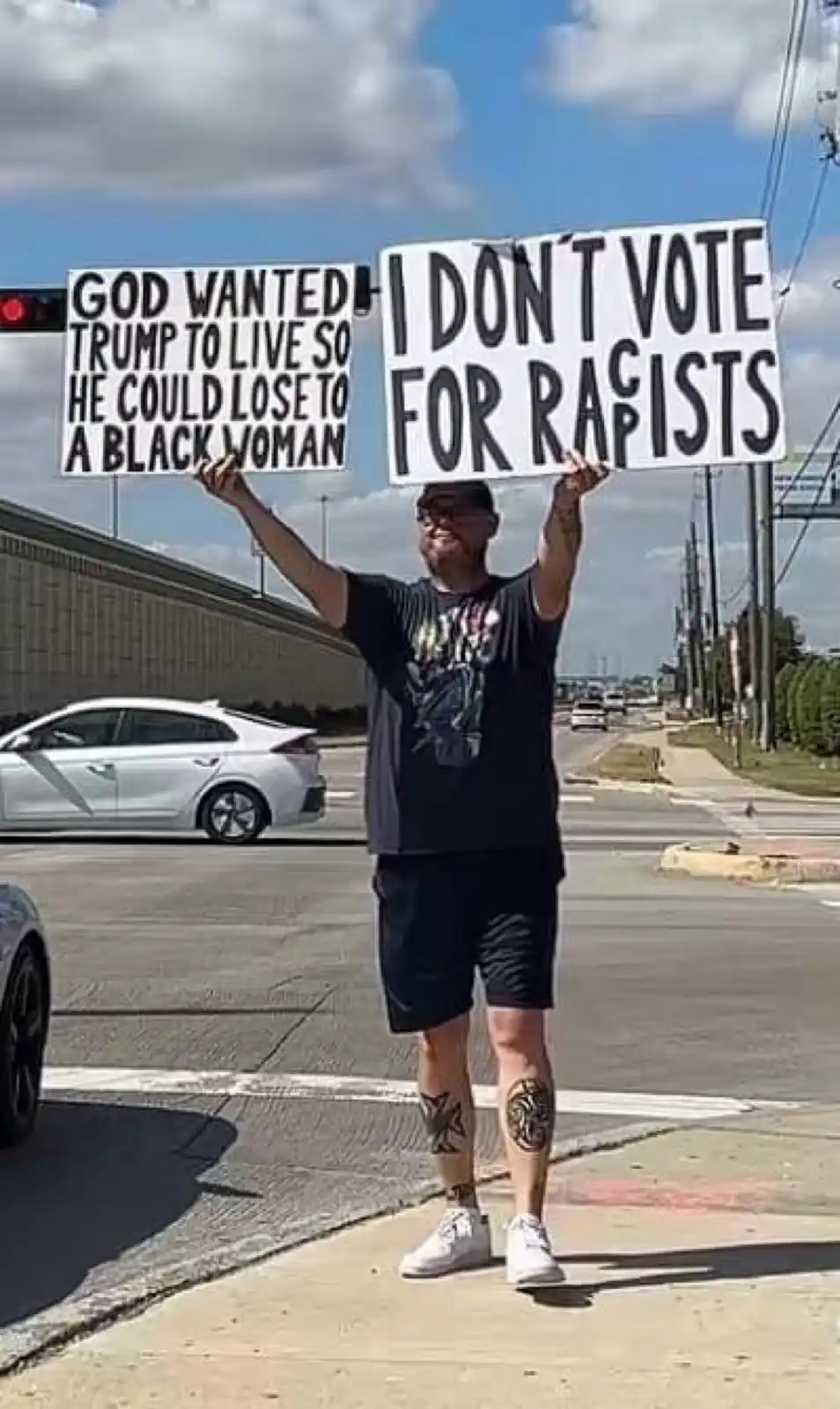 A man stands on a sidewalk at a street intersection holding up two white protest signs with black text. One sign reads 'GOD…