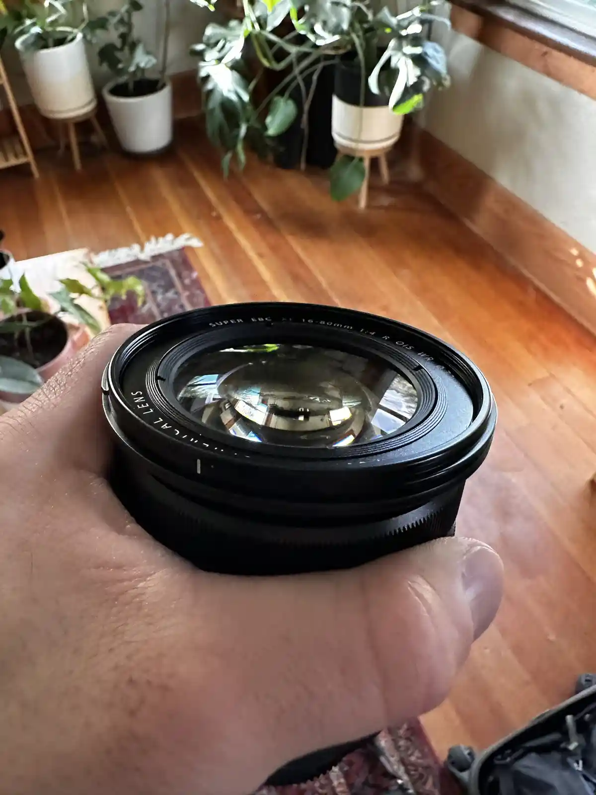 A hand holds a black camera lens with visible text 'SUPER BBC XF 16-80mm 1:4 R OIS WR' and 'MECHANICAL LENS' on its barrel…