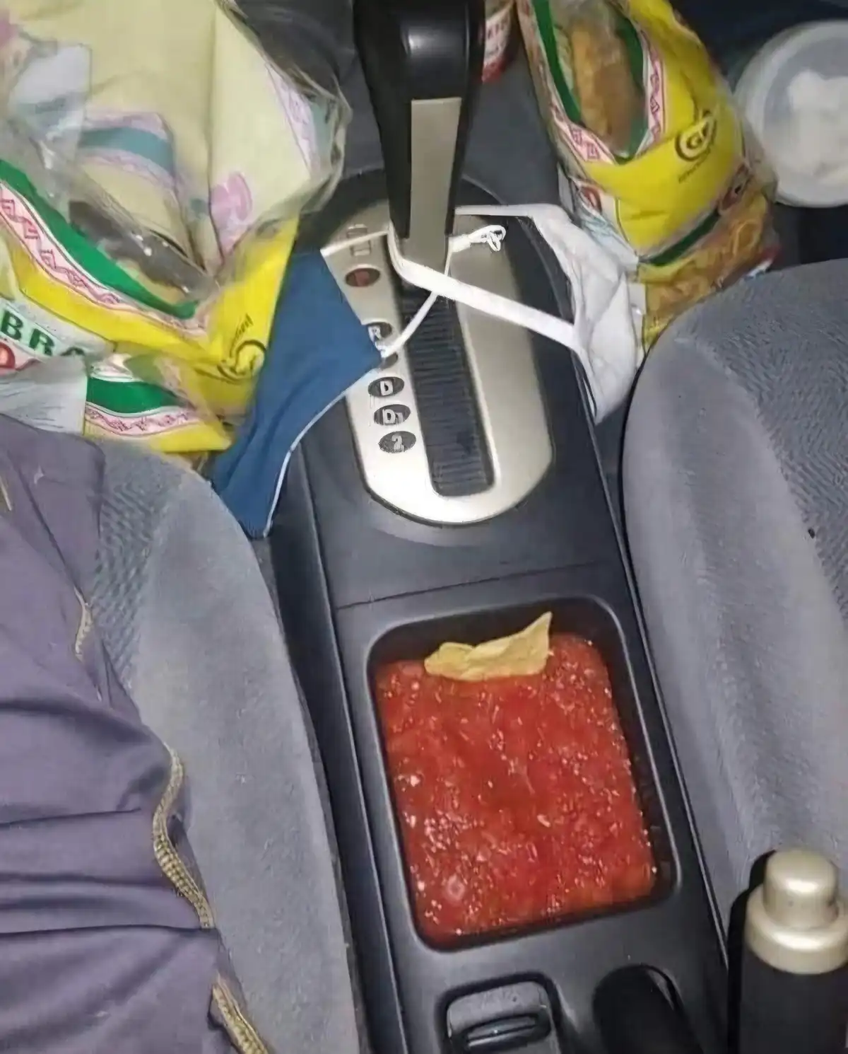 A cluttered car center console shows a messy cup holder filled with red sauce and a single chip, surrounded by bags of…