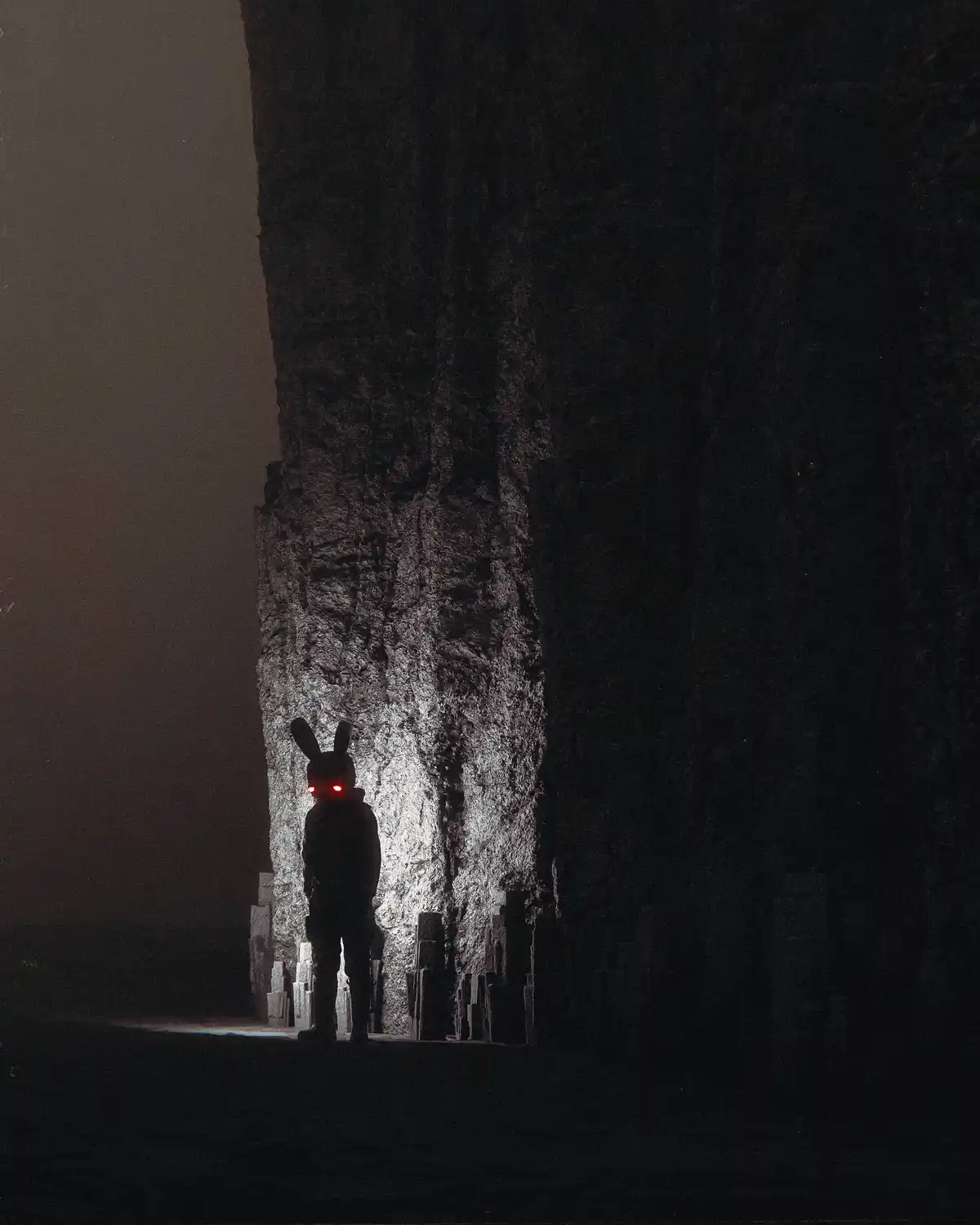 A person wearing a bunny mask with glowing red eyes stands in a dark, misty, rocky canyon. The figure is silhouetted against…