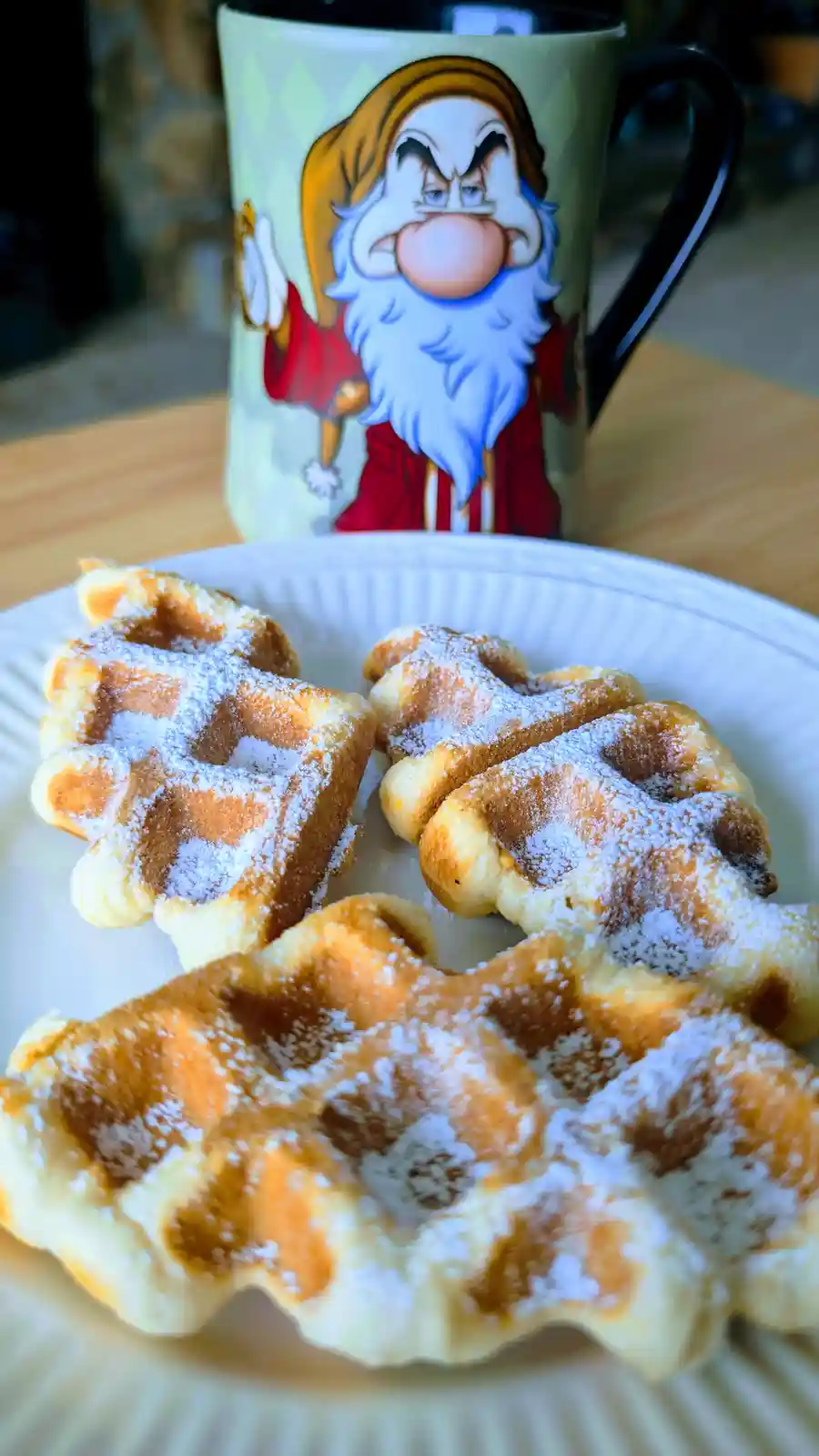 A plate of golden-brown, powdered sugar-dusted waffles sits in the foreground, with a mug featuring the angry face of Grumpy…