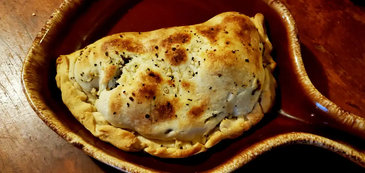 a golden-brown, baked calzone sits in a rustic brown ceramic dish on a wooden table, sprinkled with herbs and spices. the…