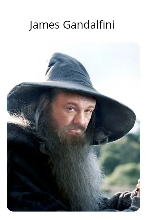 A man with a long gray beard and a large pointed wizard hat, dressed in dark robes, looks directly at the camera with a…