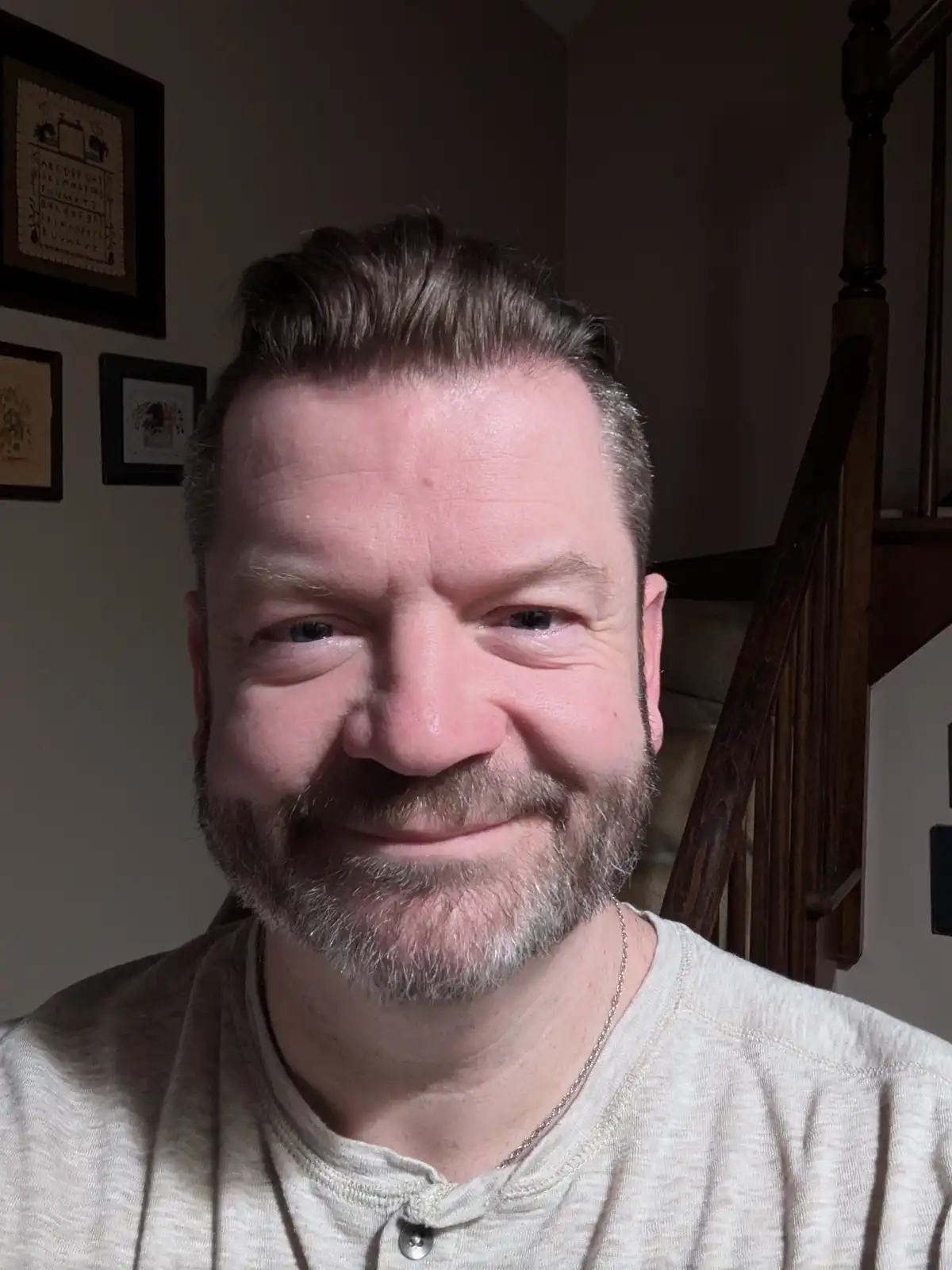 A close-up selfie of a middle-aged man with a graying beard and hair, wearing a light-colored henley shirt and a thin chain…