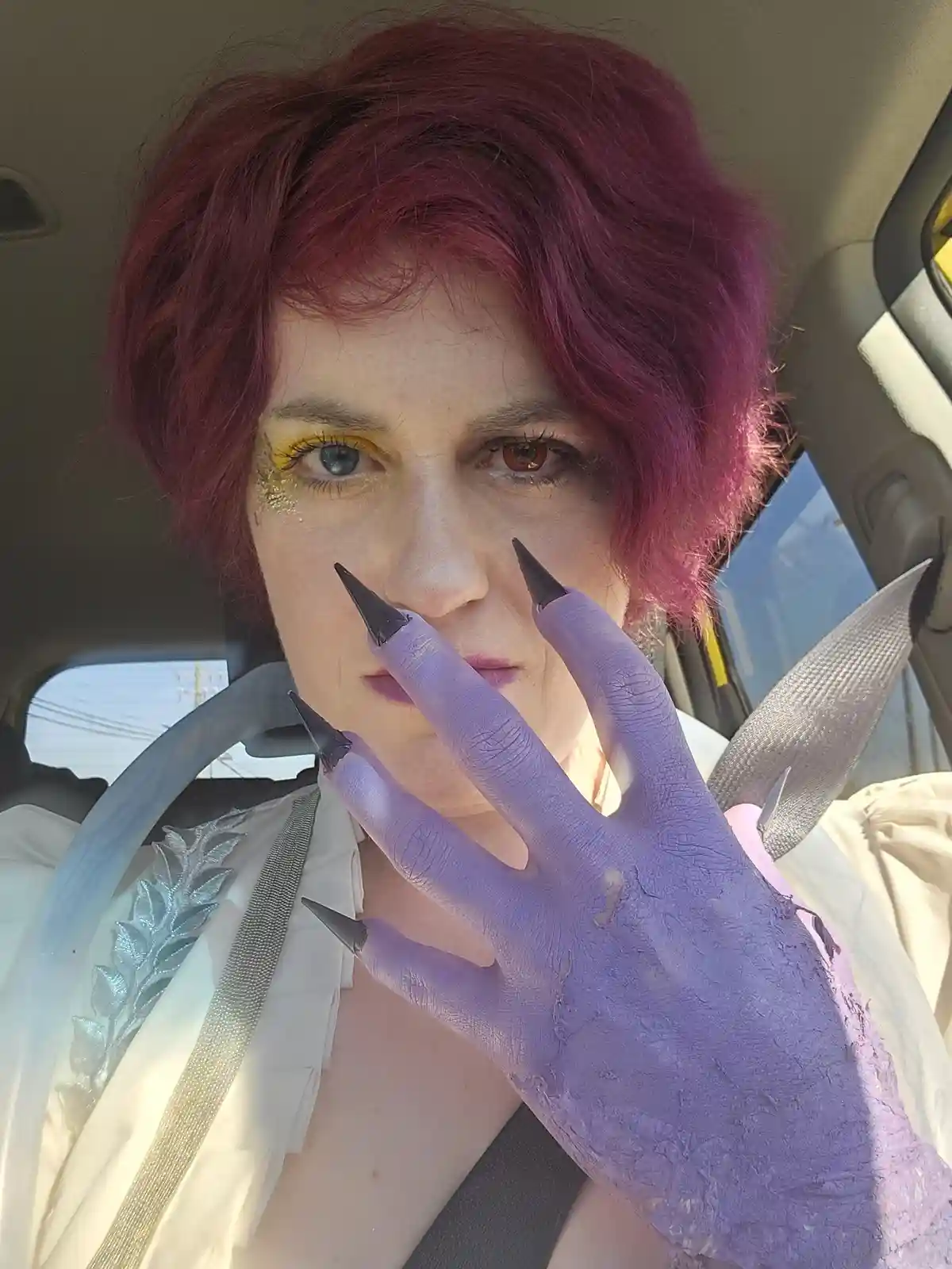 A person with short magenta hair and dramatic makeup, including yellow glitter around one eye and purple-painted hands with…