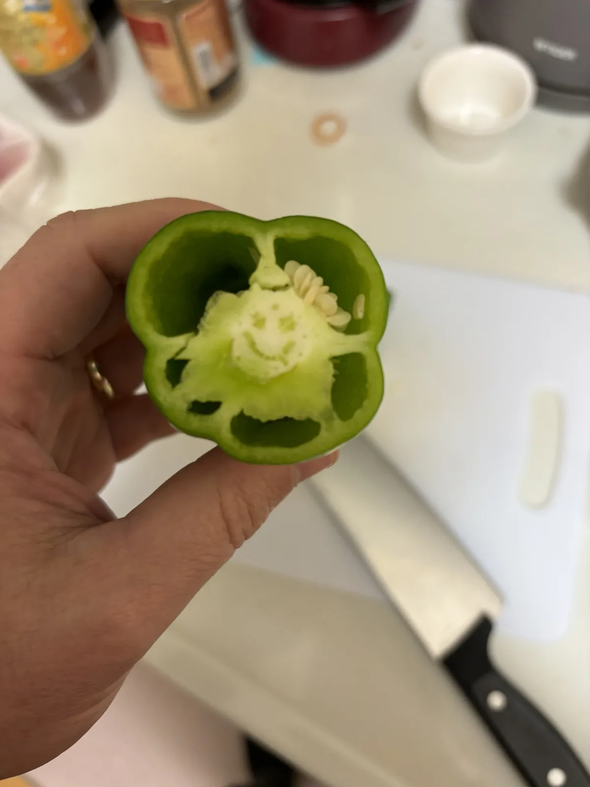 A hand holds a halved green bell pepper, revealing a natural, smiling face pattern formed by its internal seeds and…
