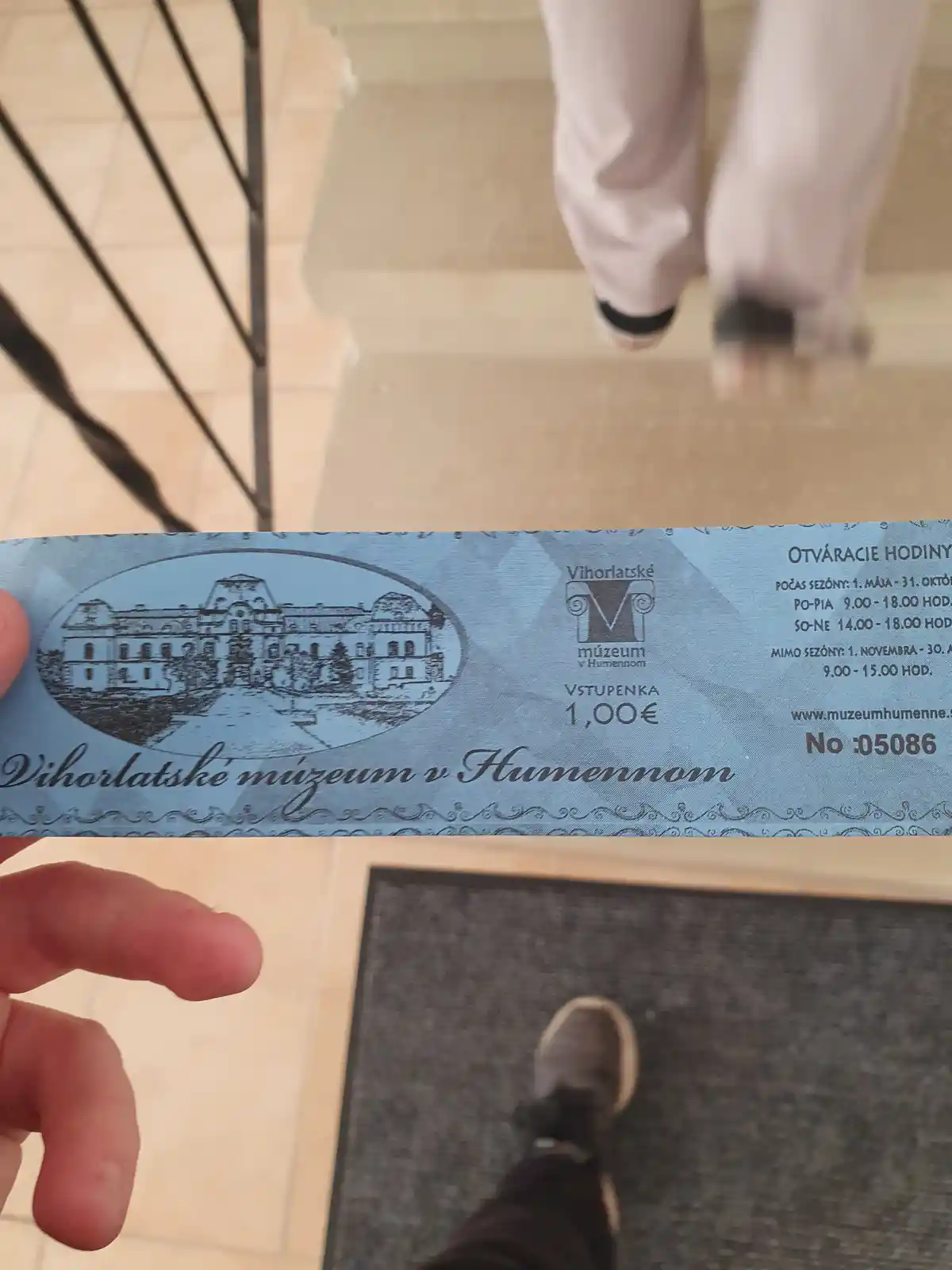 A person holds up a blue museum ticket for the Vihorlatske muzeum v Humennom, which costs 1,00€, while standing on a tiled…