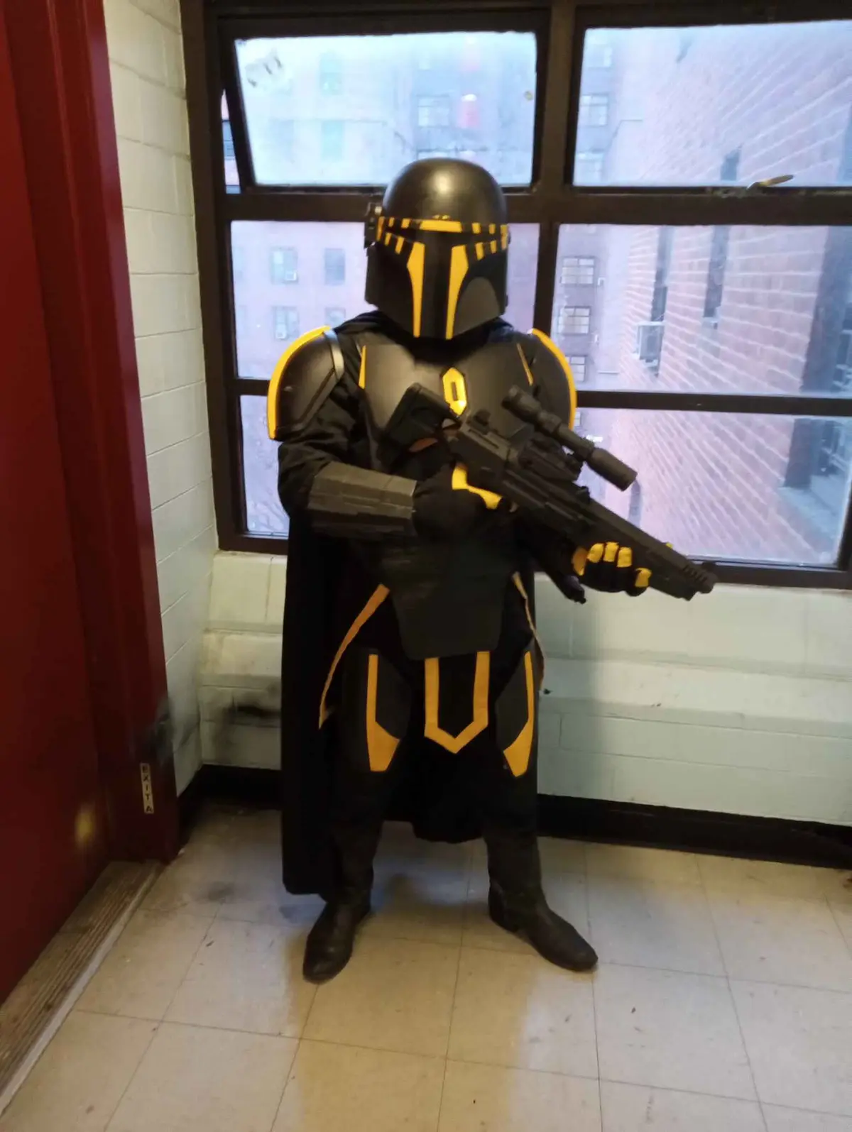 A person in a full-body Mandalorian-style costume with black and yellow armor, helmet, and cape, holding a large sci-fi…