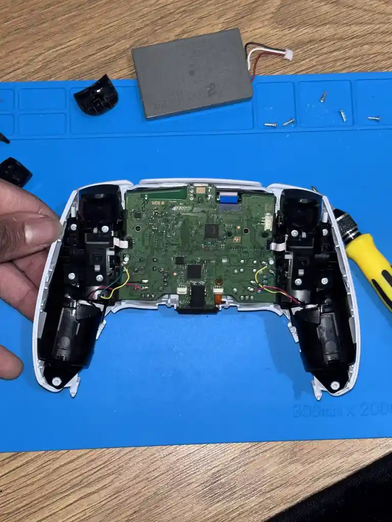 A disassembled white video game controller lies on a blue anti-static mat, revealing its green circuit board, internal…