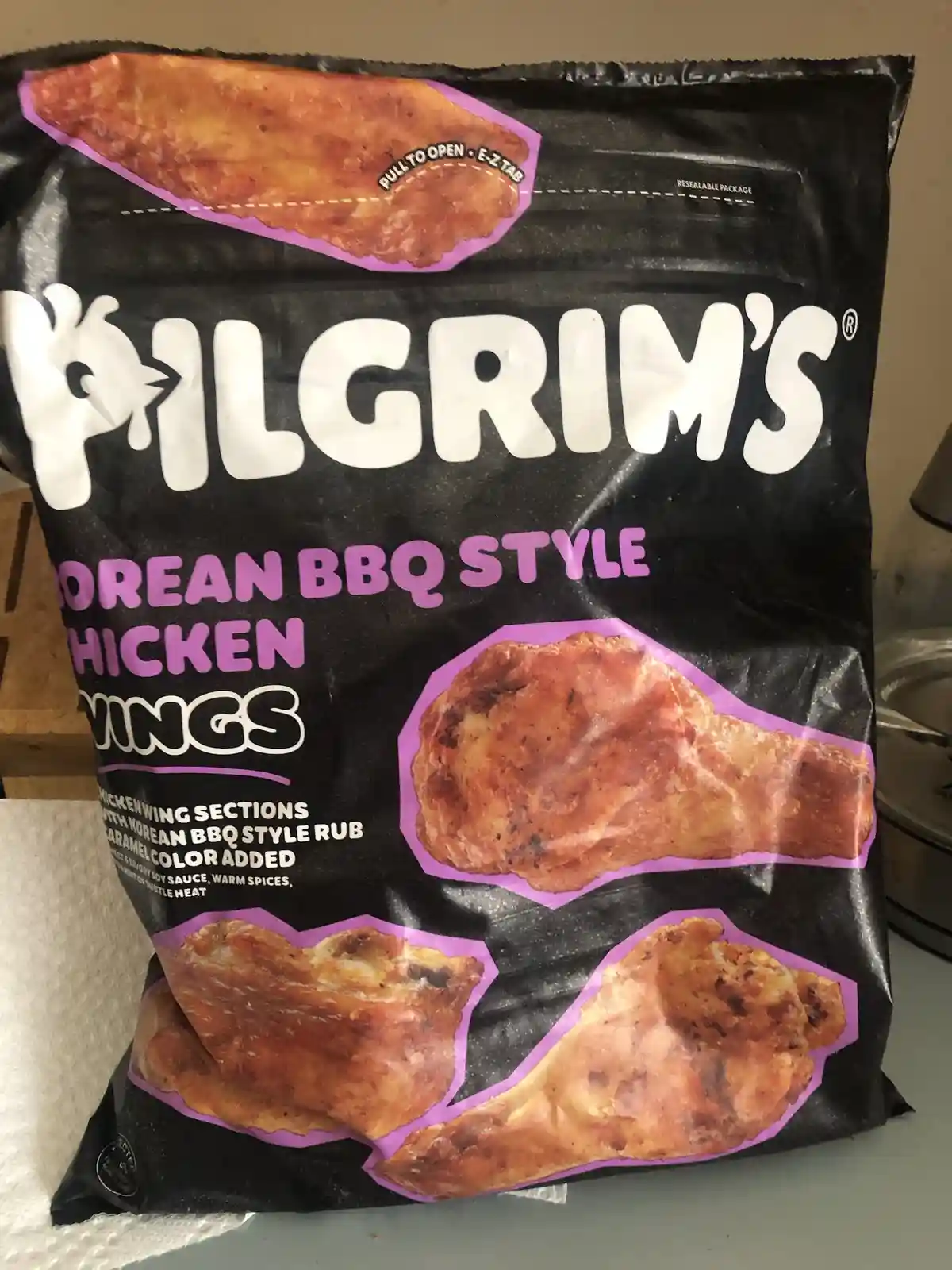 A black bag of Pilgrim's Korean BBQ Style Chicken Wings shows images of the seasoned chicken pieces. The text on the bag…