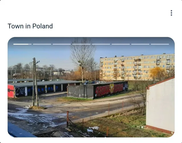 An outdoor photo of a town in Poland showing a large, weathered apartment building, a low concrete wall with red graffiti, a…