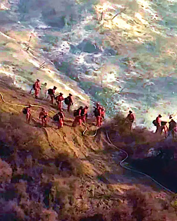 A group of firefighters in red uniforms are walking in a line along a steep, rocky hillside next to a body of water, with a…