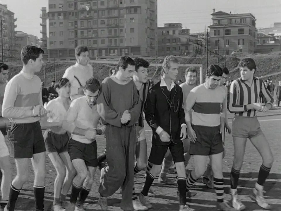 A black-and-white photo shows a group of men in vintage soccer uniforms walking across a field, some looking serious or…