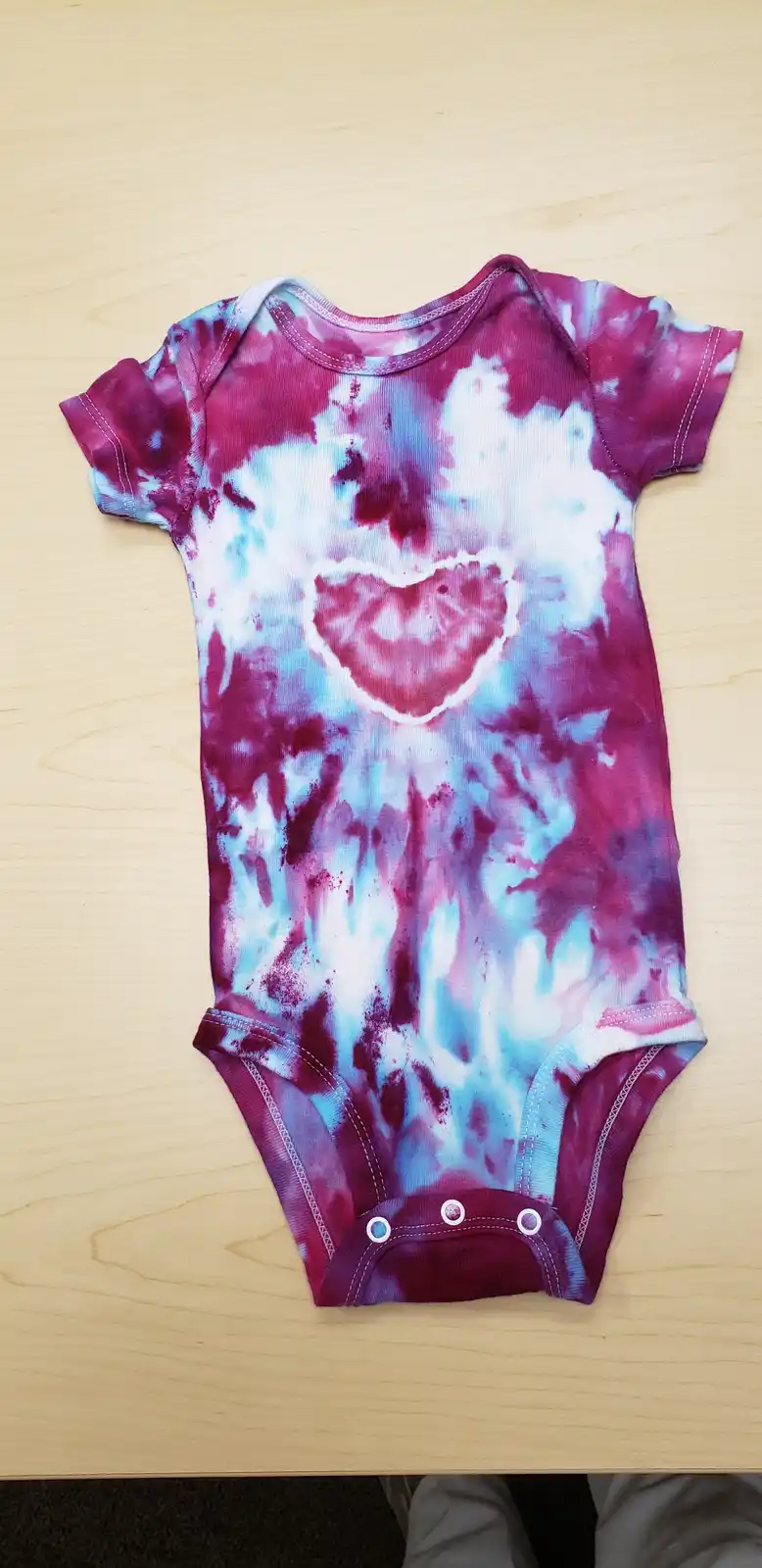 A tie-dye baby onesie with a heart-shaped design in magenta and light blue is laid flat on a light wooden surface. The…