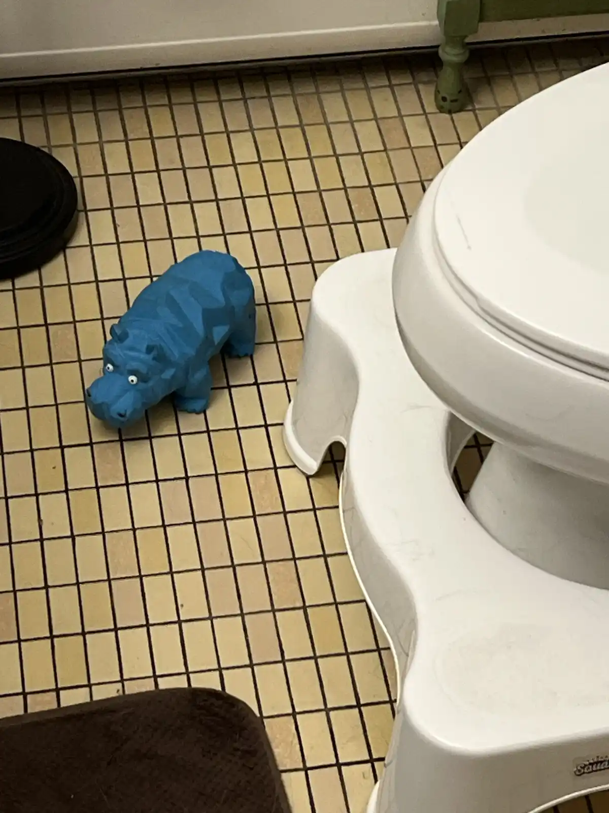 A blue, low-poly style hippo toy with wide, startled eyes sits on a beige tiled bathroom floor next to a white child's potty…