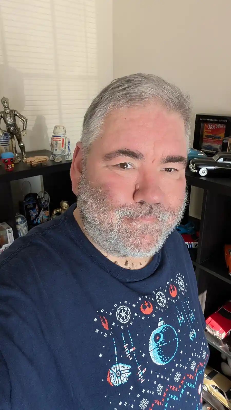 A middle-aged man with gray hair and beard wears a dark blue Star Wars-themed t-shirt featuring pixelated Death Star…