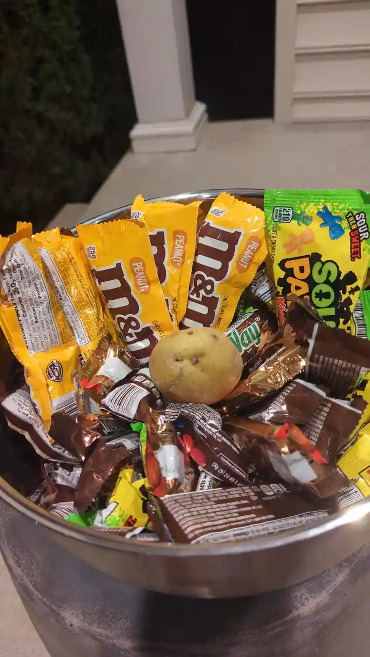 A metal bowl filled with various candy packages, including peanut M&Ms and sour patch kids, has a single potato placed…