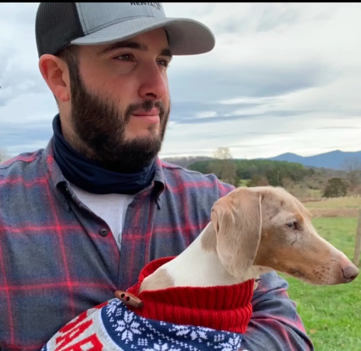 A man with a beard and a grey and black baseball cap holds a light brown and white dog wearing a festive red and blue…